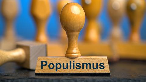 Ein Holzstempel mit der Aufschrift "Populismus" auf einer Schieferplatte mit weiteren Stempeln unscharf dahinter vor einem blauen Hintergrund. 