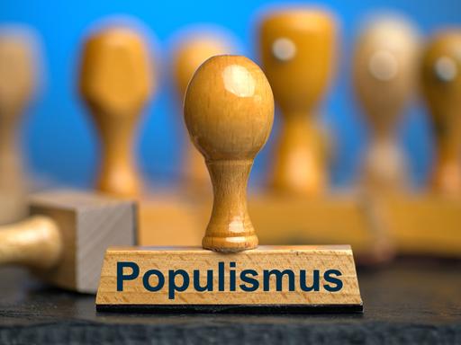 Ein Holzstempel mit der Aufschrift "Populismus" auf einer Schieferplatte mit weiteren Stempeln unscharf dahinter vor einem blauen Hintergrund. Ein Holzstempel mit der Aufschrift "Populismus" auf einer Schieferplatte mit weiteren Stempeln unscharf dahinter vor einem blauen Hintergrund.