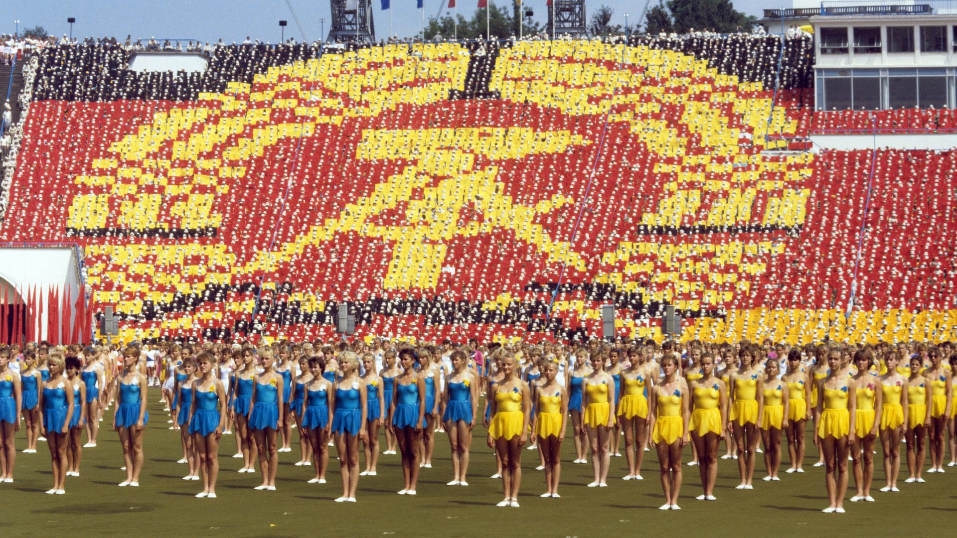 Gelb und blau gekleidete Turnerinnen stehen in Reihe beim Turn- und Sportfest in Leipzig 1987 vor einem überdimensionalen DDR-Symbol, das Teilnehmende auf den Rängen eines Sportstadions bilden.