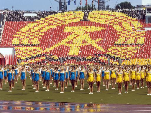 Gelb und blau gekleidete Turnerinnen stehen in Reihe beim Turn- und Sportfest in Leipzig 1987 vor einem überdimensionalen DDR-Symbol, das Teilnehmende auf den Rängen eines Sportstadions bilden.