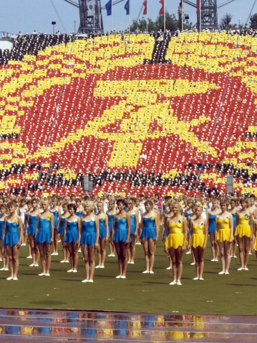 Gelb und blau gekleidete Turnerinnen stehen in Reihe beim Turn- und Sportfest in Leipzig 1987 vor einem überdimensionalen DDR-Symbol, das Teilnehmende auf den Rängen eines Sportstadions bilden.