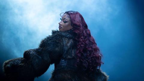 Megan Thee Stallion performt auf einer Bühne. Langes dunkelrotes Haar wallt über ihren Rücken und sie trägt einen Pelz.