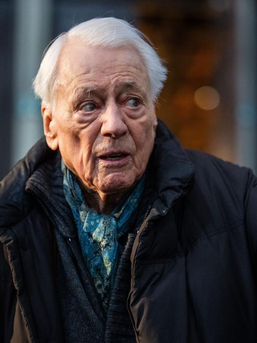 Alexander Kluge steht in einem Museum und deutet auf etwas außerhalb des Bildes