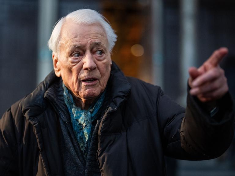 Alexander Kluge steht in einem Museum und deutet auf etwas außerhalb des Bildes