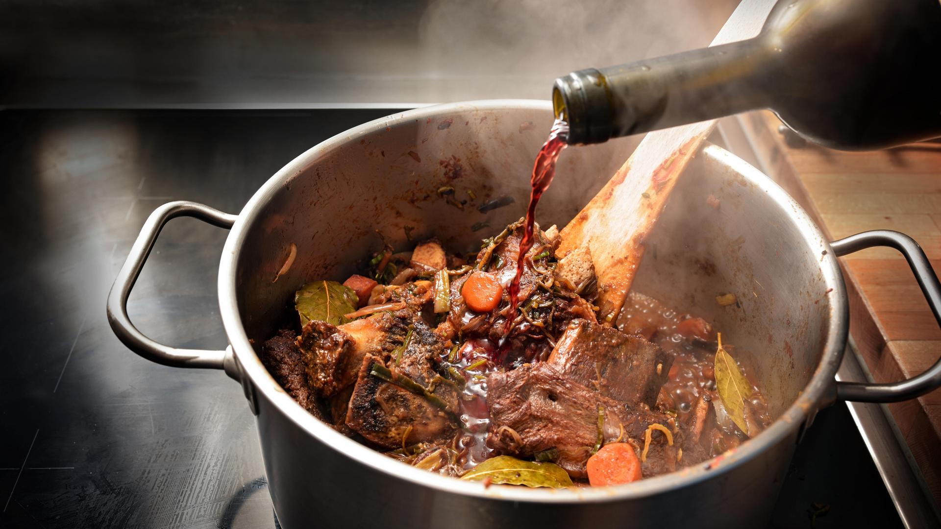 Rotwein wird in einen dampfenden Edelstahltopf gegossen, um die gebratenen Zutaten wie Knochen, Fleisch und Gemüse abzulöschen