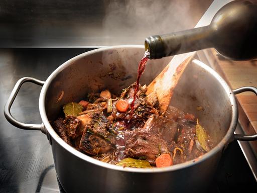 Rotwein wird in einen dampfenden Edelstahltopf gegossen, um die gebratenen Zutaten wie Knochen, Fleisch und Gemüse abzulöschen