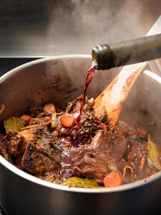 Rotwein wird in einen dampfenden Edelstahltopf gegossen, um die gebratenen Zutaten wie Knochen, Fleisch und Gemüse abzulöschen