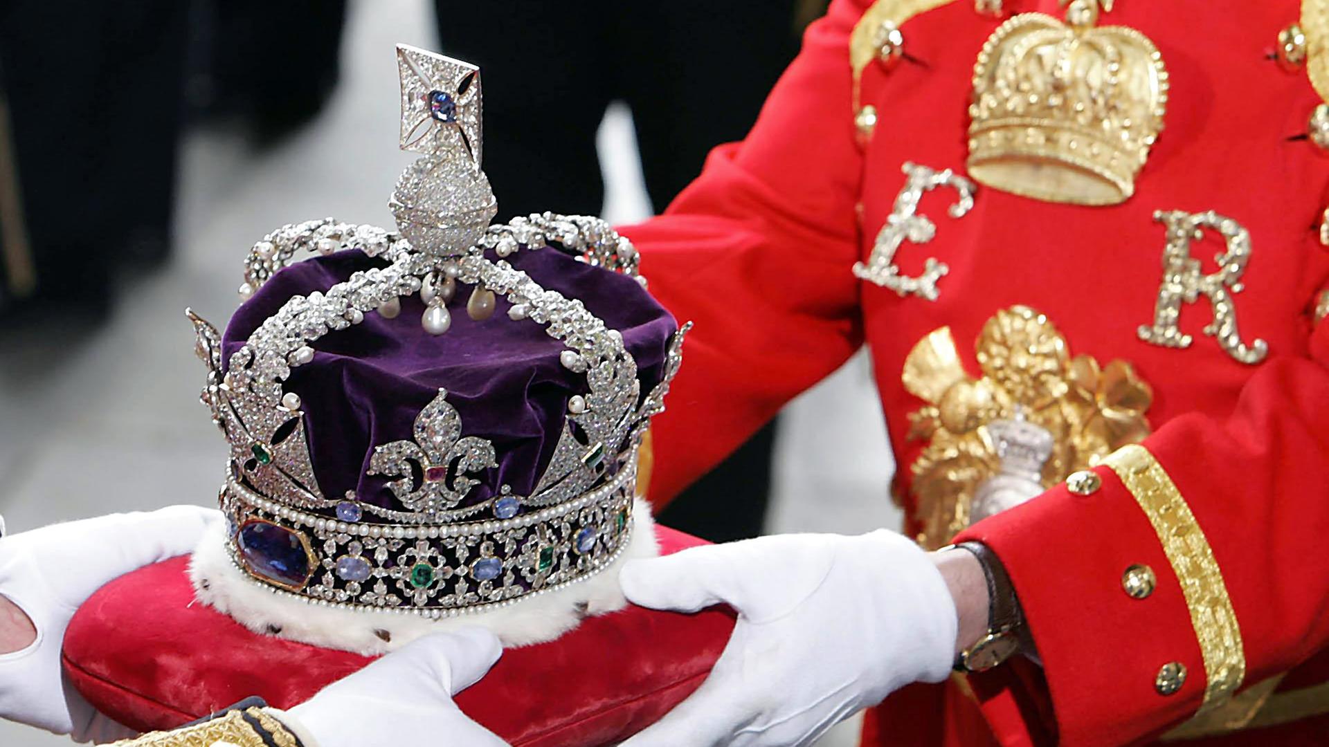 Die Krone der Königen Elizabeth II. ruht auf einem roten Kissen, während zwei Personen in Galauniform und weißen Handschuhen das Kissen halten.