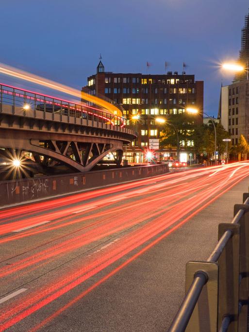 Nacht in einer Stadt: Auf einer von Laternen gesäumten Straße verlaufen leuchtenden roten Linien von fahrenden Autos, links davon eine Brücke mit einer Lichtlinie von einer fahrenden U-Bahn, im Hintergrund ein Haus mit beleuchteten Fenstern.