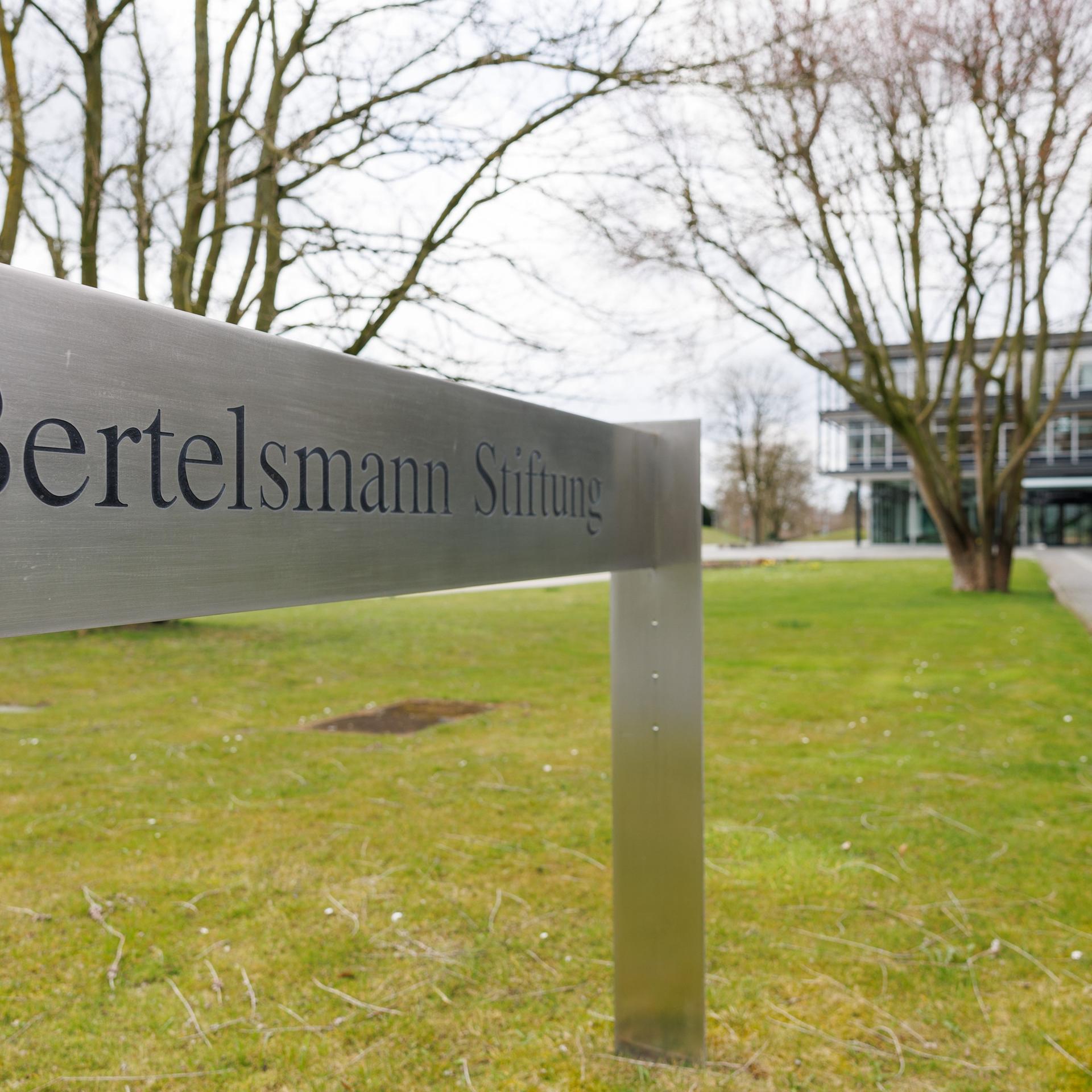 Blick auf ein Schild "Bertelsmann Stiftung" - im Hintergrund ist verschwommen ein Gebäudekomplex zu sehen