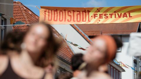 Banner über einer Gasse in der der Alttstadt, auf dem der Schriftzug Rudolstadt Festival zu lesen ist. Davor sind zwei junge Frauen verschwommen wahrzunehmen, die auf der Straße flanieren.