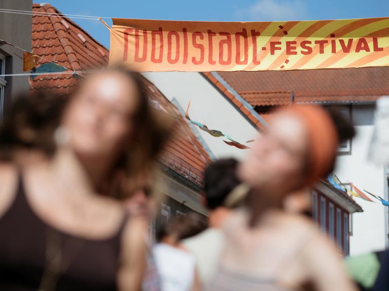 Banner über einer Gasse in der der Alttstadt, auf dem der Schriftzug Rudolstadt Festival zu lesen ist. Davor sind zwei junge Frauen verschwommen wahrzunehmen, die auf der Straße flanieren.