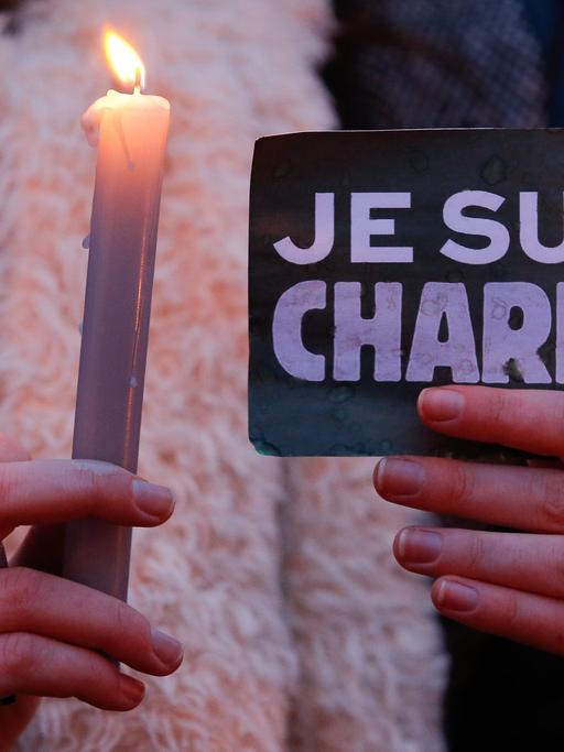 Eine Frau hält eine Kerze, einen Kugelschreiber und eine Schild mit der AUfschrift "Je suis Charlie"