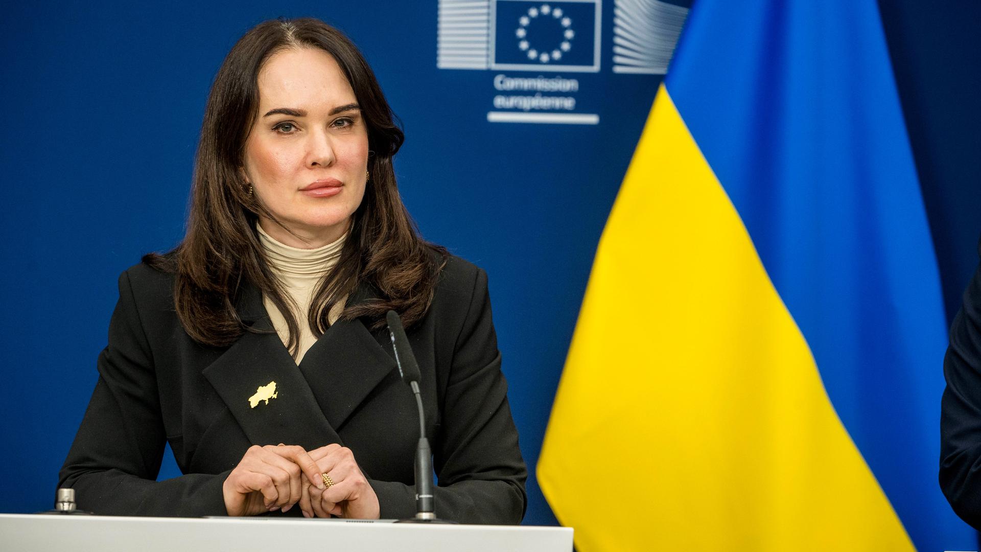 Irina Mudra sitzt in einer Pressekonferenz vor einem Mikrofon. Links neben ihr ist die ukrainische Fahne zu sehen.