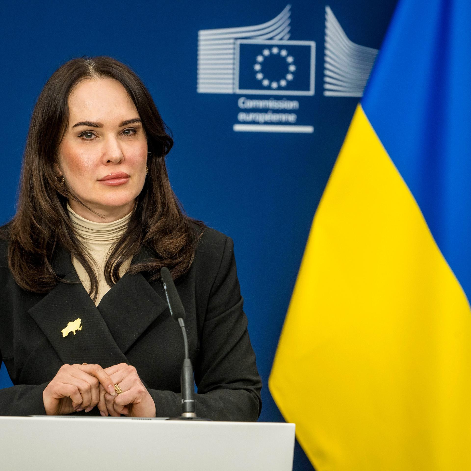 Irina Mudra sitzt in einer Pressekonferenz vor einem Mikrofon. Links neben ihr ist die ukrainische Fahne zu sehen.