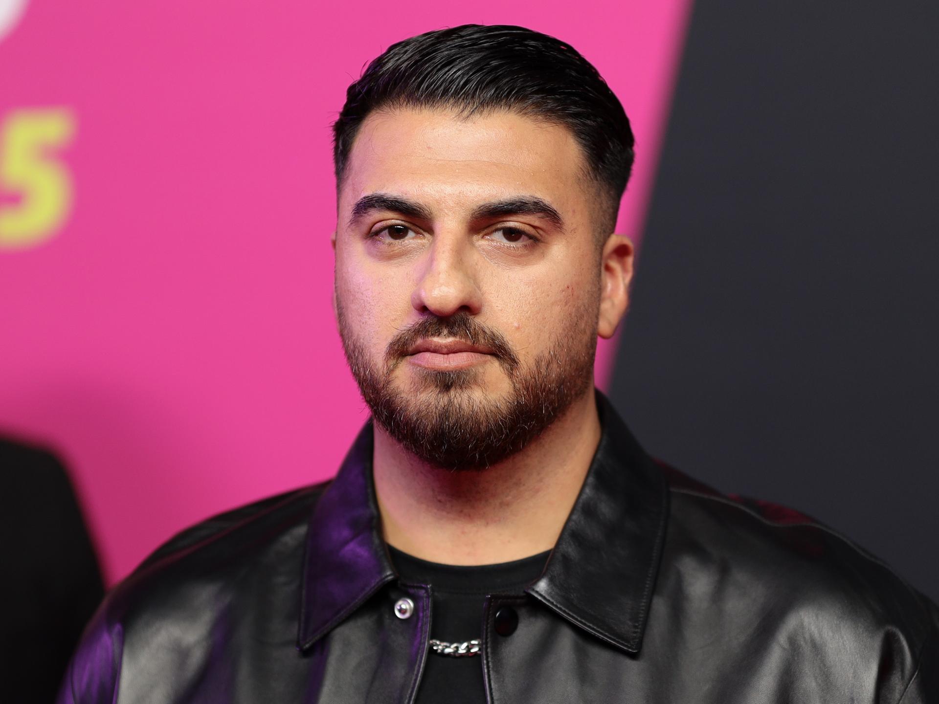 Tahsim Durgun steht auf einer Bühne vor einem pinkfarbenen Hintergrund, blickt direkt in die Kamera und trägt eine schwarze Jacke. Tahsim Durgun steht auf einer Bühne vor einem pinkfarbenen Hintergrund, blickt direkt in die Kamera und trägt eine schwarze Jacke.