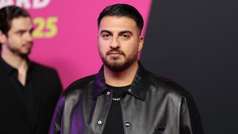 Tahsim Durgun steht auf einer Bühne vor einem pinkfarbenen Hintergrund, blickt direkt in die Kamera und trägt eine schwarze Jacke. Tahsim Durgun steht auf einer Bühne vor einem pinkfarbenen Hintergrund, blickt direkt in die Kamera und trägt eine schwarze Jacke.