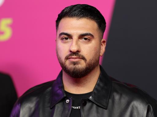 Tahsim Durgun steht auf einer Bühne vor einem pinkfarbenen Hintergrund, blickt direkt in die Kamera und trägt eine schwarze Jacke. Tahsim Durgun steht auf einer Bühne vor einem pinkfarbenen Hintergrund, blickt direkt in die Kamera und trägt eine schwarze Jacke.