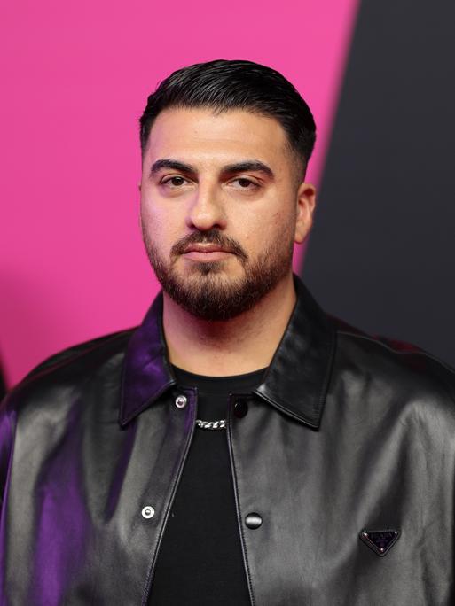 Tahsim Durgun steht auf einer Bühne vor einem pinkfarbenen Hintergrund, blickt direkt in die Kamera und trägt eine schwarze Jacke. Tahsim Durgun steht auf einer Bühne vor einem pinkfarbenen Hintergrund, blickt direkt in die Kamera und trägt eine schwarze Jacke.