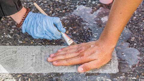 Zu sehen ist eine Handfläche, die auf Asphalt geklebt ist und durch einen Beamten mit Hilfe von einem Pinsel mit Flüssigkeit bearbeitet wird.