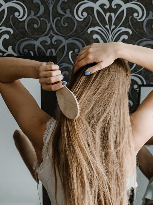 Ein ganz normaler Tag beginnt: Der Blick in den Spiegel zeigt nicht das Guten-Morgen-Gewinnergesicht. Zu sehen: Eine junge Frau vor einem Spiegel kämmt sich ihre langen Haare. 