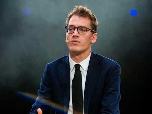 Arthur Mensch in Anzug und Krawatte, vor grün-gräulichem Hintergrund. Er trägt eine Brille und schaut in Richtung der Kamera, allerdings leicht vorbei.