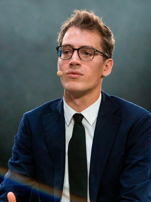 Arthur Mensch in Anzug und Krawatte, vor grün-gräulichem Hintergrund. Er trägt eine Brille und schaut in Richtung der Kamera, allerdings leicht vorbei.