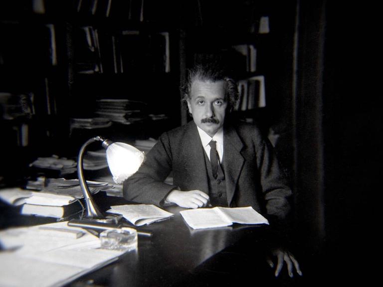 Physiker Albert Einstein sitzt an einem Schreibtisch. Vor ihm liegt ein geöffnetes Notizbuch.