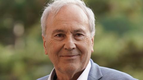 Ein Mann mit weißgrauem Haar blickt freundlich schmunzelnd in die Kamera. Er trägt ein weißes Hemd und blaugrau meliertes Jackett. Im Hintergrund ist verschwommenes Grün zu sehen.