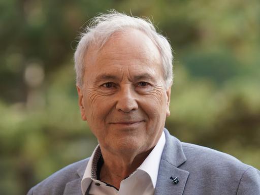 Ein Mann mit weißgrauem Haar blickt freundlich schmunzelnd in die Kamera. Er trägt ein weißes Hemd und blaugrau meliertes Jackett. Im Hintergrund ist verschwommenes Grün zu sehen.