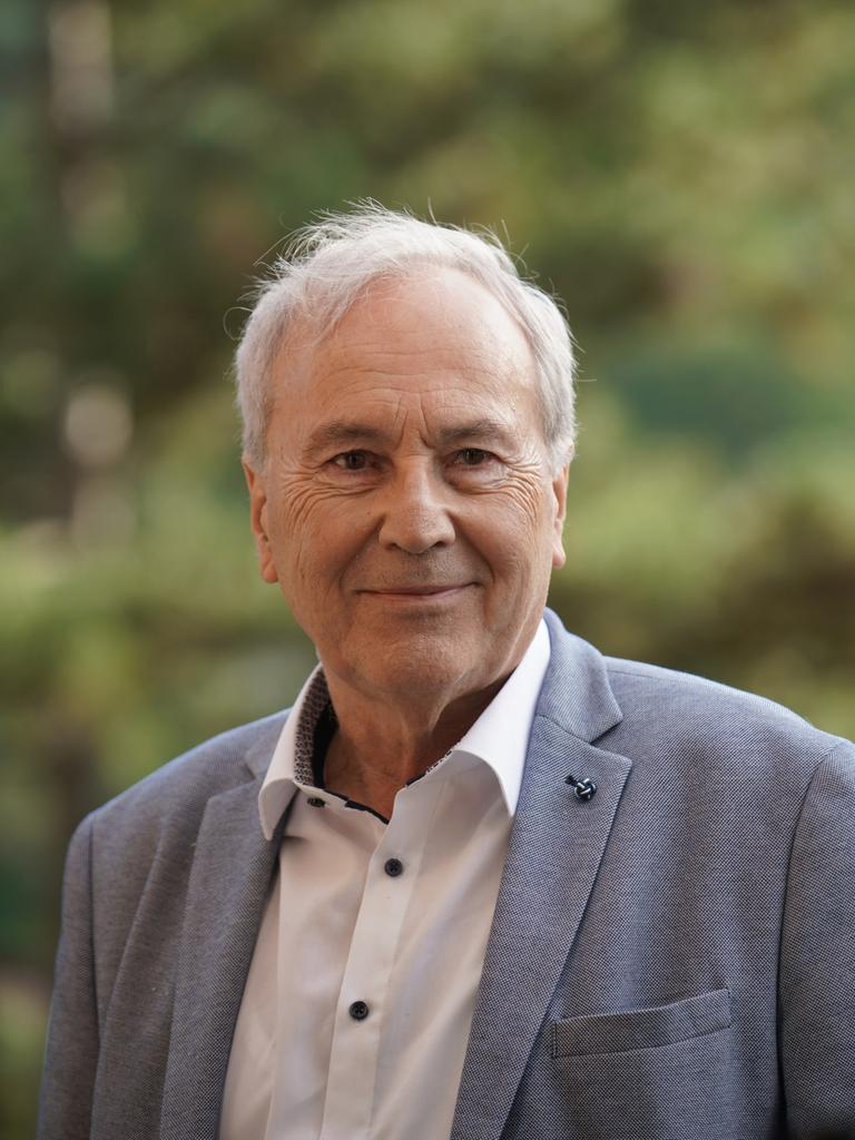 Ein Mann mit weißgrauem Haar blickt freundlich schmunzelnd in die Kamera. Er trägt ein weißes Hemd und blaugrau meliertes Jackett. Im Hintergrund ist verschwommenes Grün zu sehen.