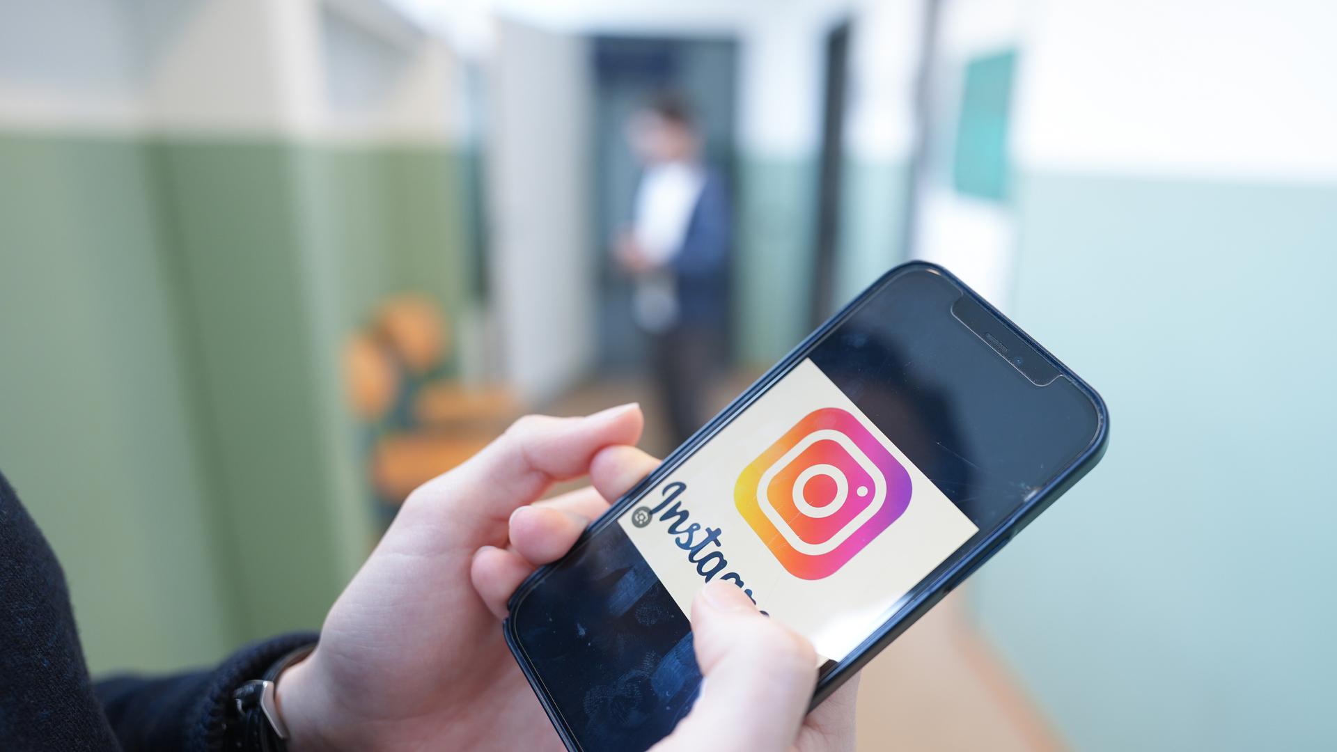 Ein Kind hält in einem Schulflur ein Smartphone in der Hand, auf dem das Logo der Social-Media-Plattform Instagram zu sehen ist.
