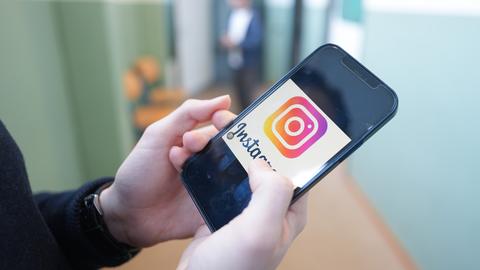 Ein Kind hält in einem Schulflur ein Smartphone in der Hand, auf dem das Logo der Social-Media-Plattform Instagram zu sehen ist.