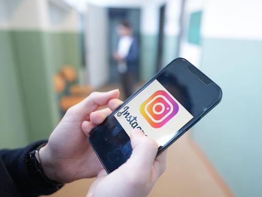 Ein Kind hält in einem Schulflur ein Smartphone in der Hand, auf dem das Logo der Social-Media-Plattform Instagram zu sehen ist.