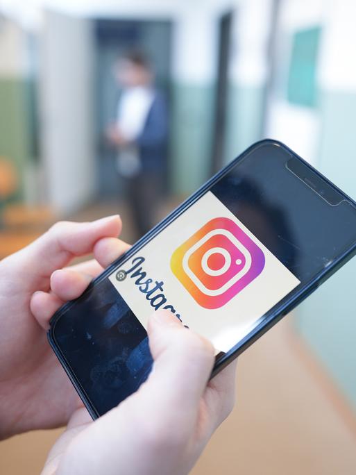 Ein Kind hält in einem Schulflur ein Smartphone in der Hand, auf dem das Logo der Social-Media-Plattform Instagram zu sehen ist.