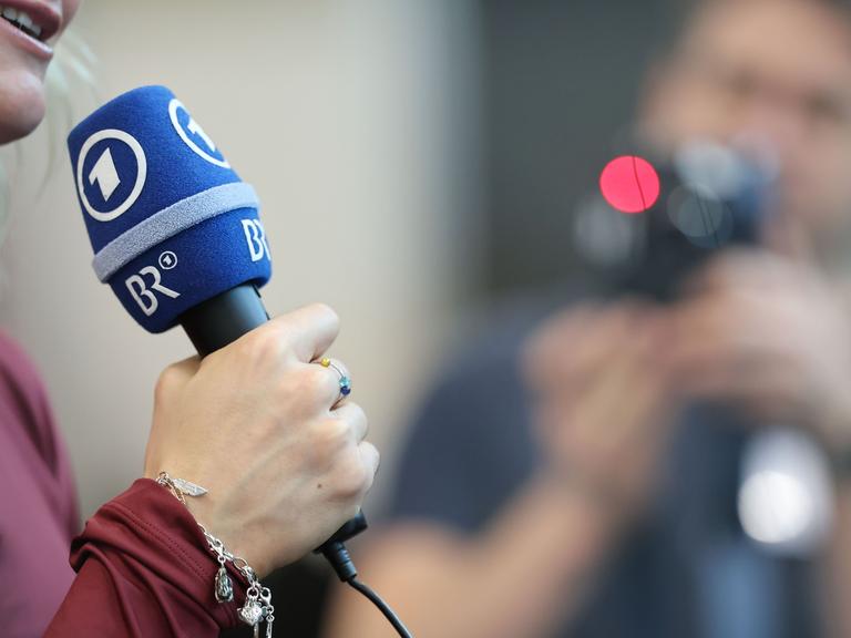 Eine Frau hält ein Mikrofon des ARD und des BR (Bayerischer Rundfunk) in der Hand und spricht, im Hintergrund ist, etwas unscharf, ein Mann mit einer Kamera in der Hand zu erkennen.