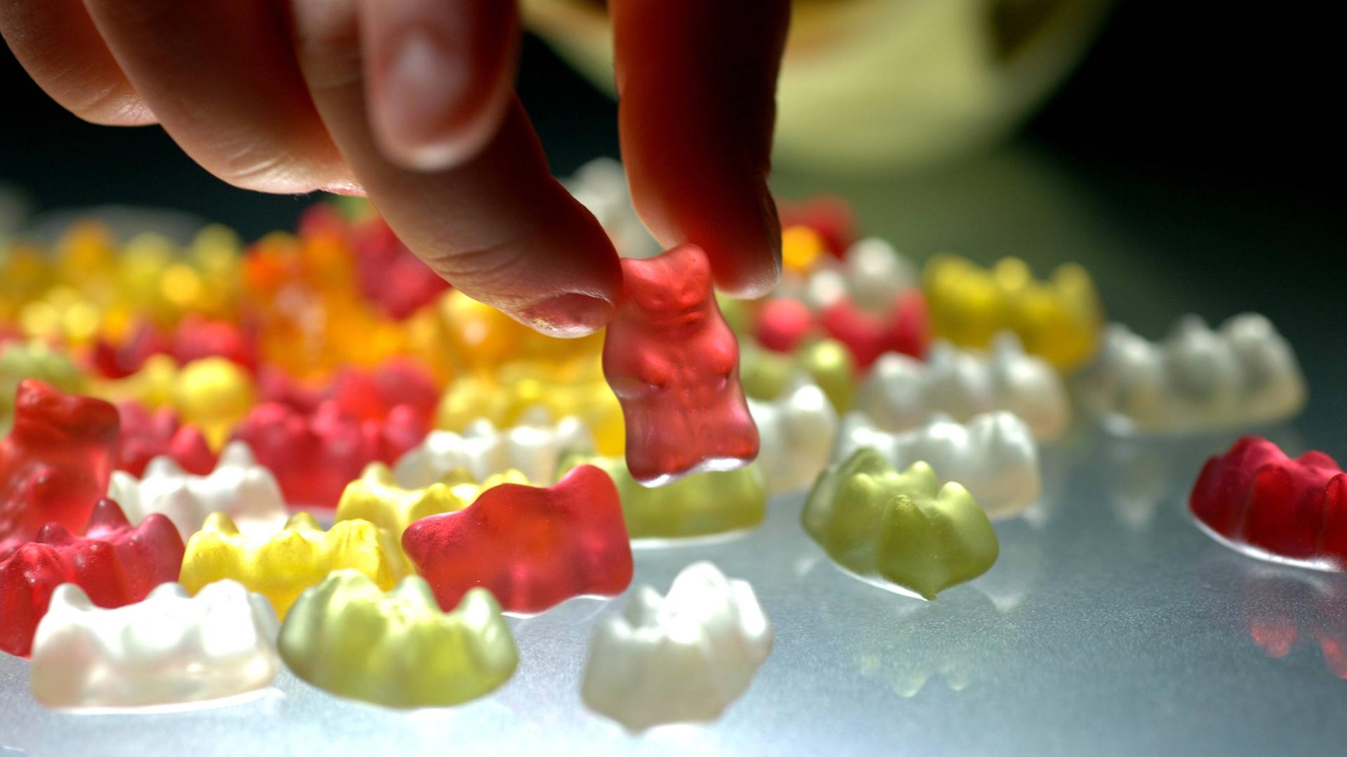 Kinderhand greift nach roten Gummibärchen, auf dem Tisch liegen weitere bunte Gummibärchen.