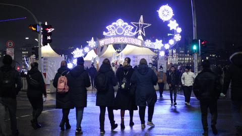 Mehrere Menschen gehen in Richtung eines Weihnachtsmarkts, dort steht auf einem Schild "Weißer Zauber"