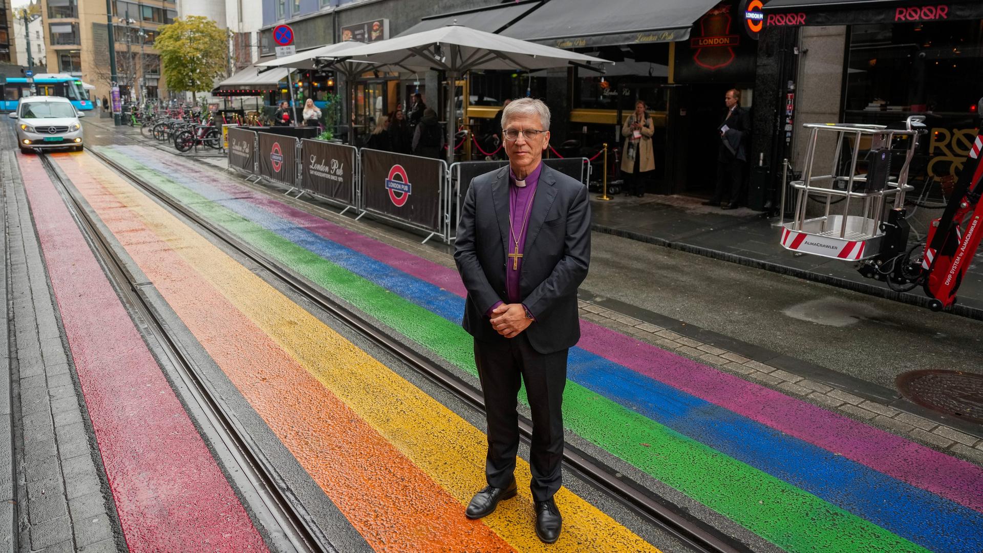 Der Bischof steht mit vor dem Körper verschränkten Händen auf der Straße in Regenbogenfarben. Dahinter der Club. Der Bischof steht mit vor dem Körper verschränkten Händen auf der Straße in Regenbogenfarben. Dahinter der Club.
