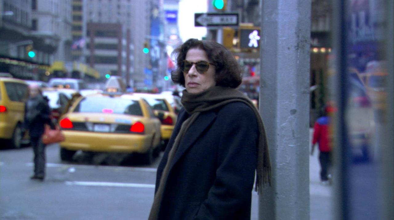 Fran Lebowitz steht in cooler Pose und mit Sonnenbrille an einer New Yorker Straße, auf der Taxis fahren.