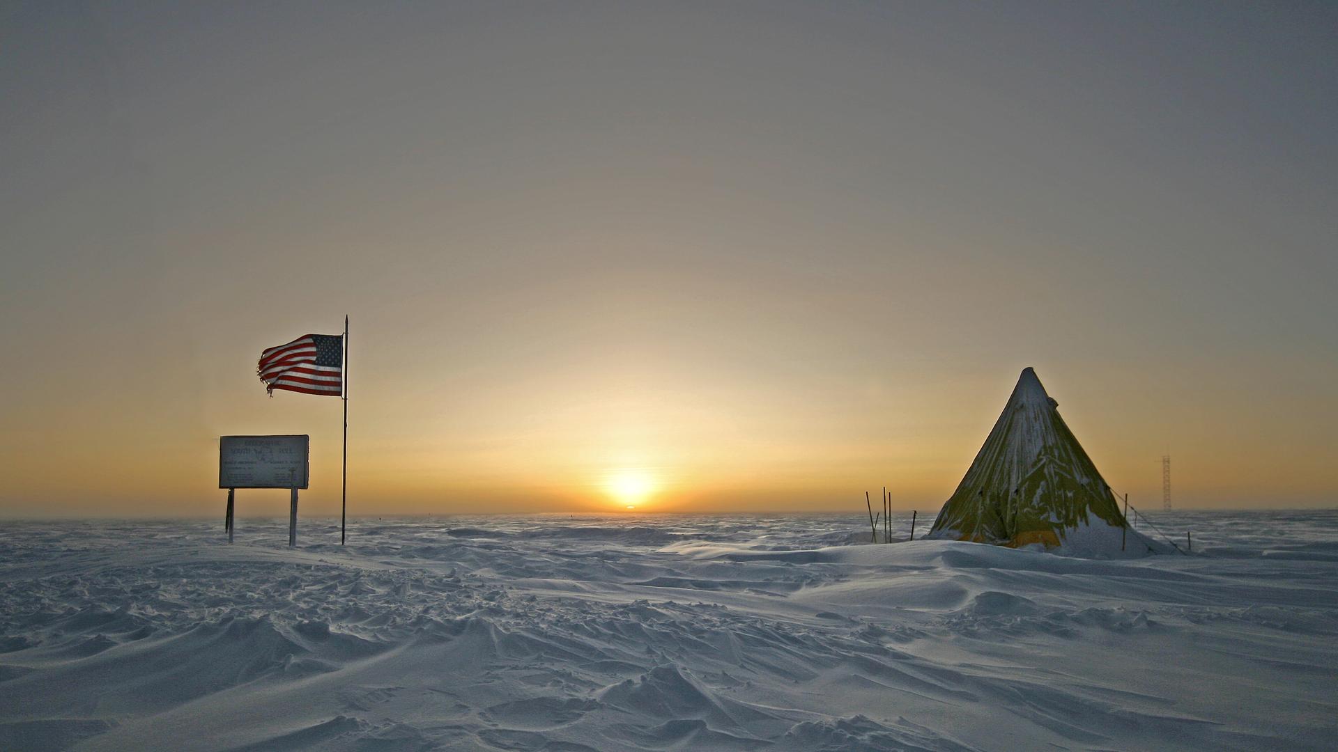 Ein Schild, eine US-Flagge und ein Zelt stehen im Eis, die Sonne ist über dem Horizont zu sehen.