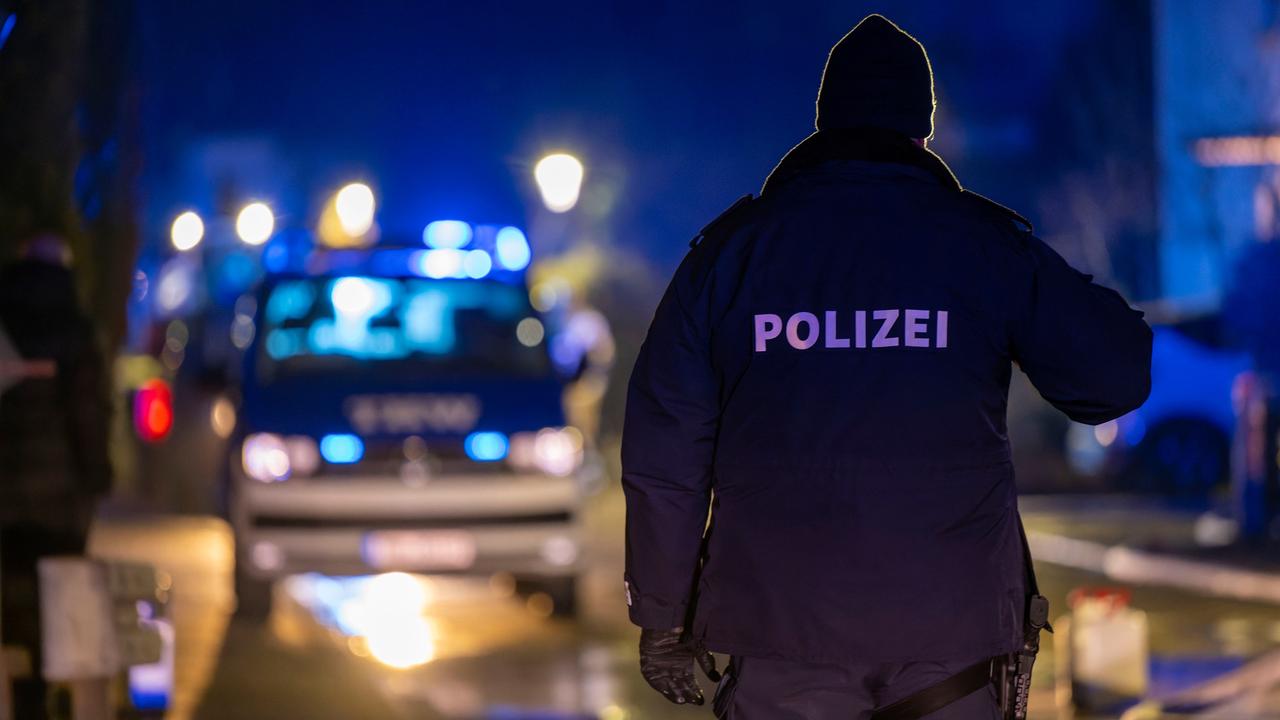 Ein Polizist steht im Dunkeln auf einer Straße mit dem Rücken zum Betrachter, im Hintergrund Blaulicht