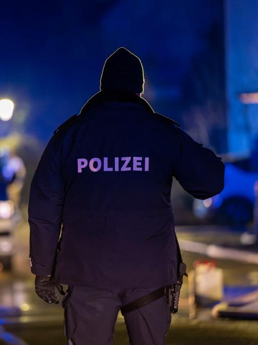 Ein Polizist steht im Dunkeln auf einer Straße mit dem Rücken zum Betrachter, im Hintergrund Blaulicht