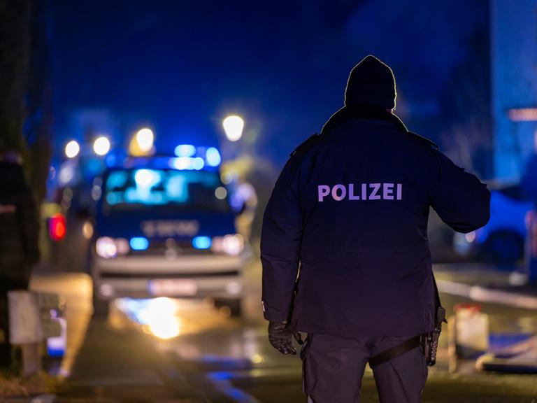 Ein Polizist steht im Dunkeln auf einer Straße mit dem Rücken zum Betrachter, im Hintergrund Blaulicht