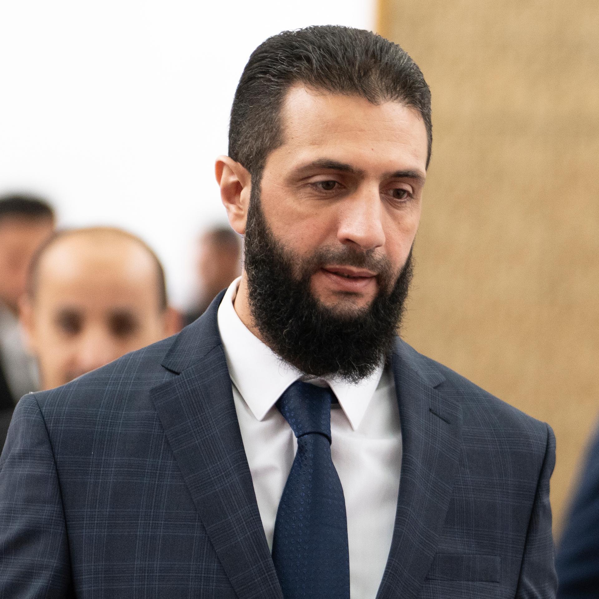 Al-Scharaa geht mit ernstem Blick einen Gang entlang. Er trägt einen schwarzen Vollbart und ein dunkles Jackett mit Krawatte. Hinter ihm unscharf weitere Personen