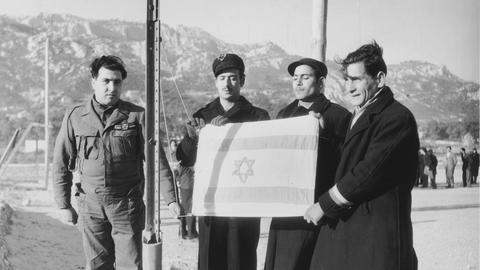 Eine Gruppe von drei jüdischen Männern (Displaced Persons) mit israelischer Fahne vor ihrer Abreise nach Palästina neben einem Soldaten (um 1948).