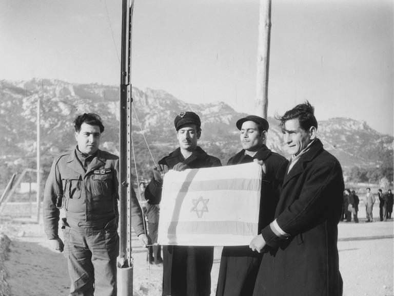Eine Gruppe von drei jüdischen Männern (Displaced Persons) mit israelischer Fahne vor ihrer Abreise nach Palästina neben einem Soldaten (um 1948).