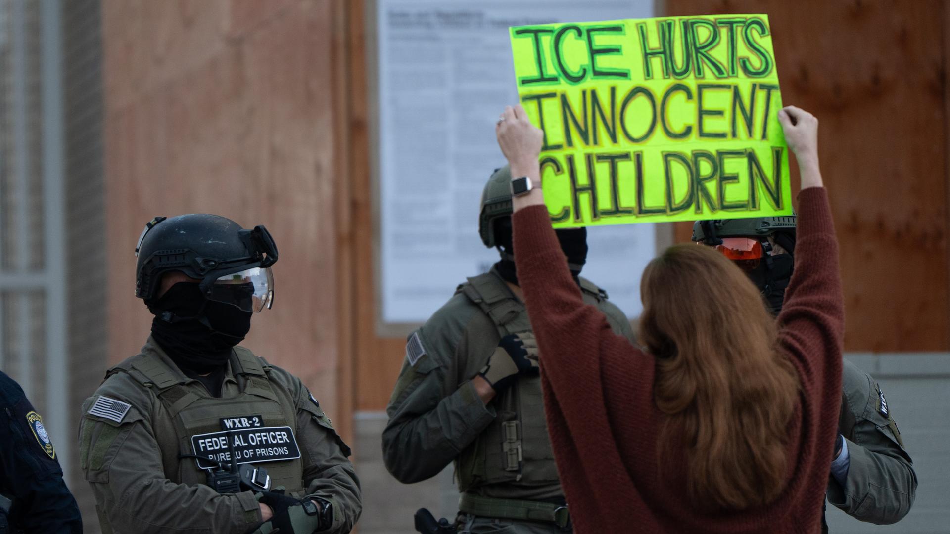 Eine Demomstrantin steht vor Männern in Uniform und hält ein Schild mit der Aufschrift "ICE hurts innocent children" - auf Deutsch: "ICE verletzt unschuldige Kinder" in die Höhe. Eine Demomstrantin steht vor Männern in Uniform und hält ein Schild mit der Aufschrift "ICE hurts innocent children" - auf Deutsch: "ICE verletzt unschuldige Kinder" in die Höhe.