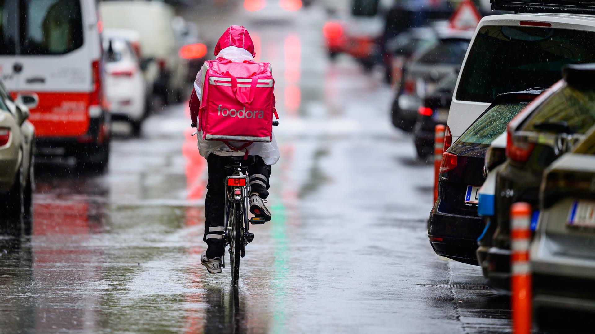 Das Bild zeigt einen Essenslieferanten mit nassem Rucksack, der in der Großstadt zwischen Autos mit einem Fahrrad durch eine regennasse Straße fährt.