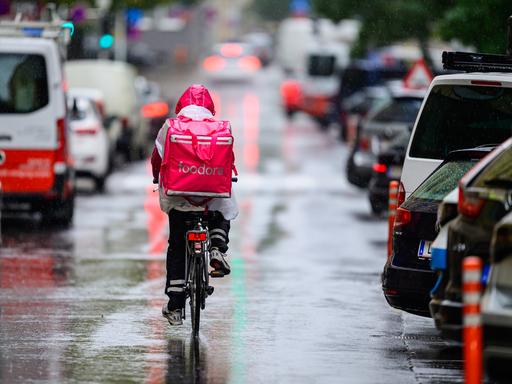Das Bild zeigt einen Essenslieferanten mit nassem Rucksack, der in der Großstadt zwischen Autos mit einem Fahrrad durch eine regennasse Straße fährt.